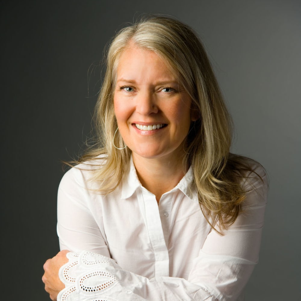 A blonde woman in a white buttoned shirt is crossing her arms and smiling.