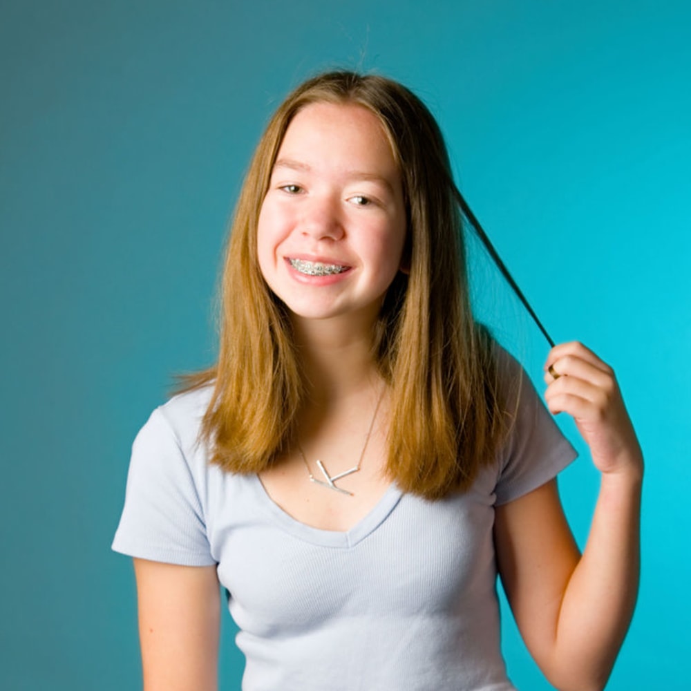 A girl with braces is smiling at the camera and playing with her hair.