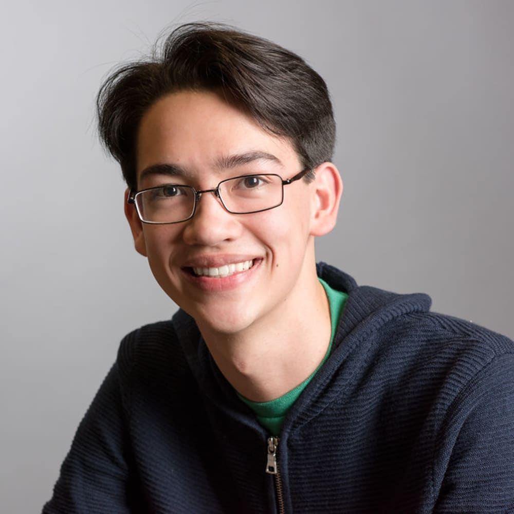 A teenage boy wearing a black sweater and glasses smiles.